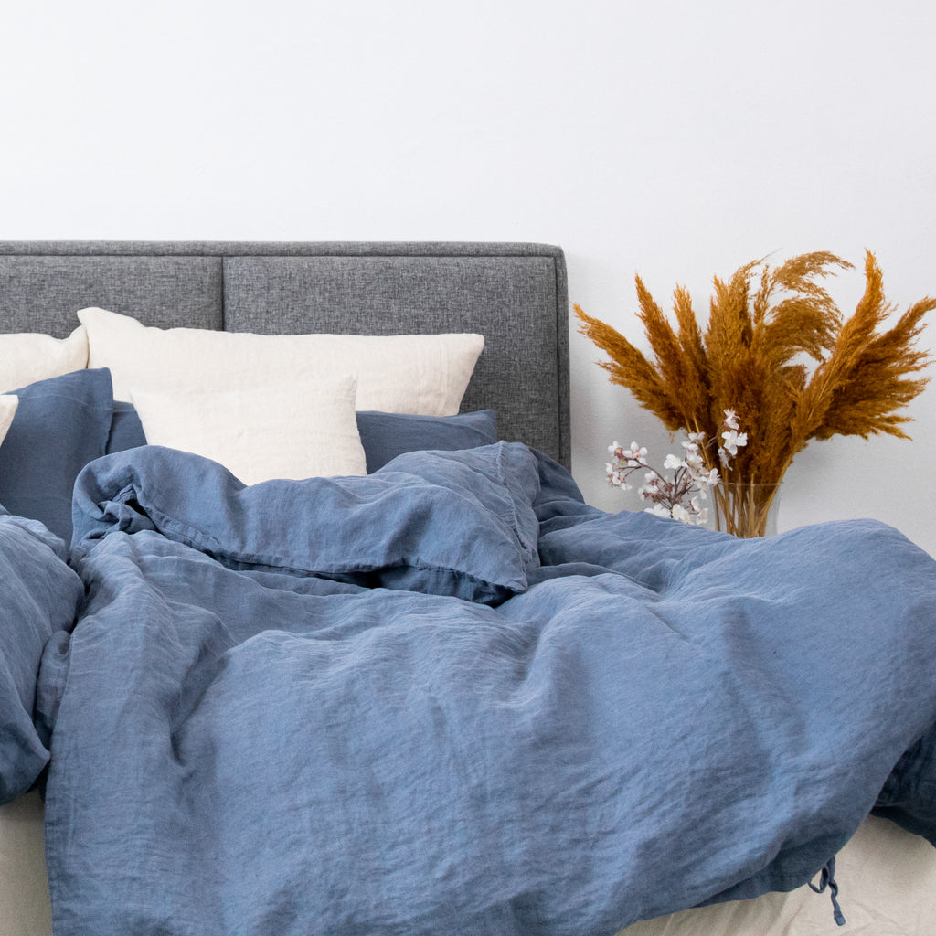 Leinen-Deckenbezug Atlantikblau auf einem Bett mit grauem Kopfteil und bunten Pflanzen im Hintergrund.