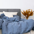 Leinen-Deckenbezug Atlantikblau auf einem Bett mit grauem Kopfteil und bunten Pflanzen im Hintergrund.